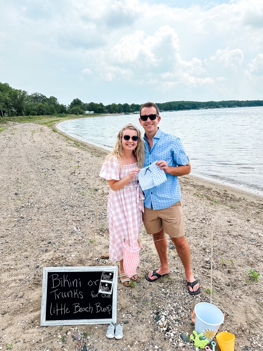 man and woman holding baby boy swim trunks on beach at gender reveal